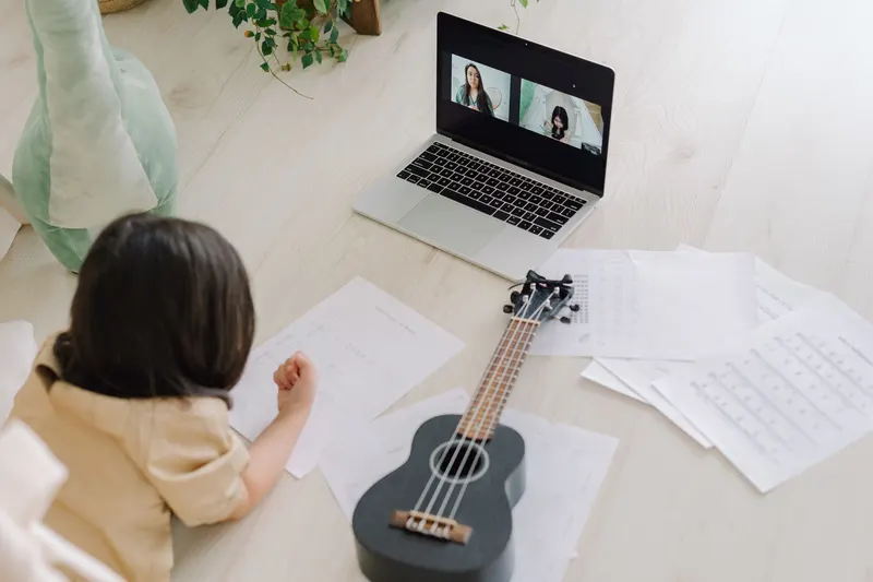 Child with a ukulele and music sheets in front of a laptop video call