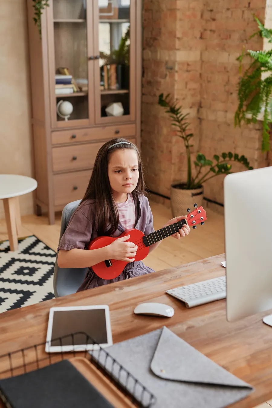 Girl playing a red ukulele at her desk in front of a computer