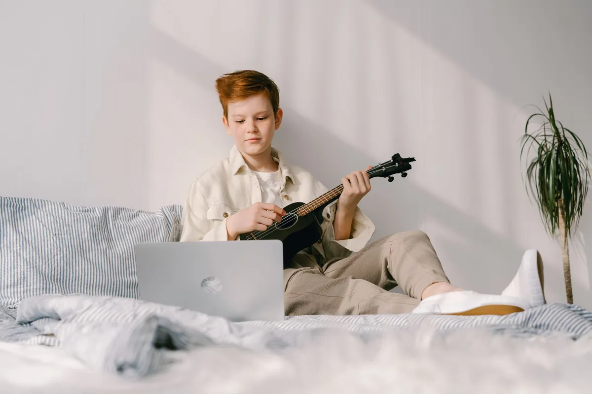 Boy playing a ukulele on his bed with a laptop in front of him
